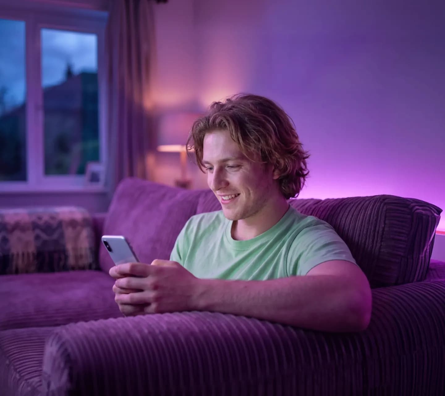 Fiatal férfi a kanapén ülve mosolyogva nézi a telefonját lila fénnyel megvilágított nappalijában