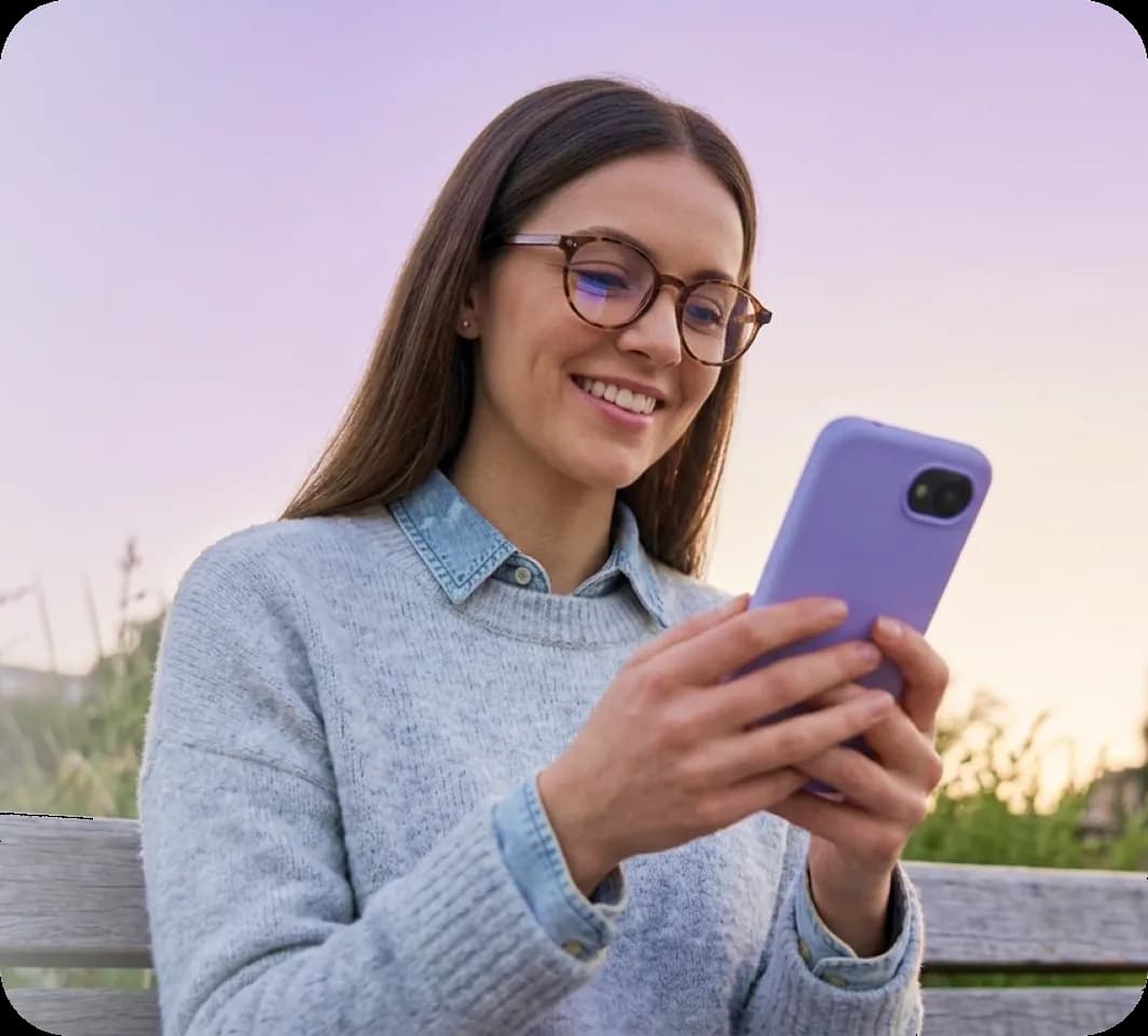 Mosolygó, szemüveges nő a szabadban, egy padon ülve a mobiltelefonját nézi
