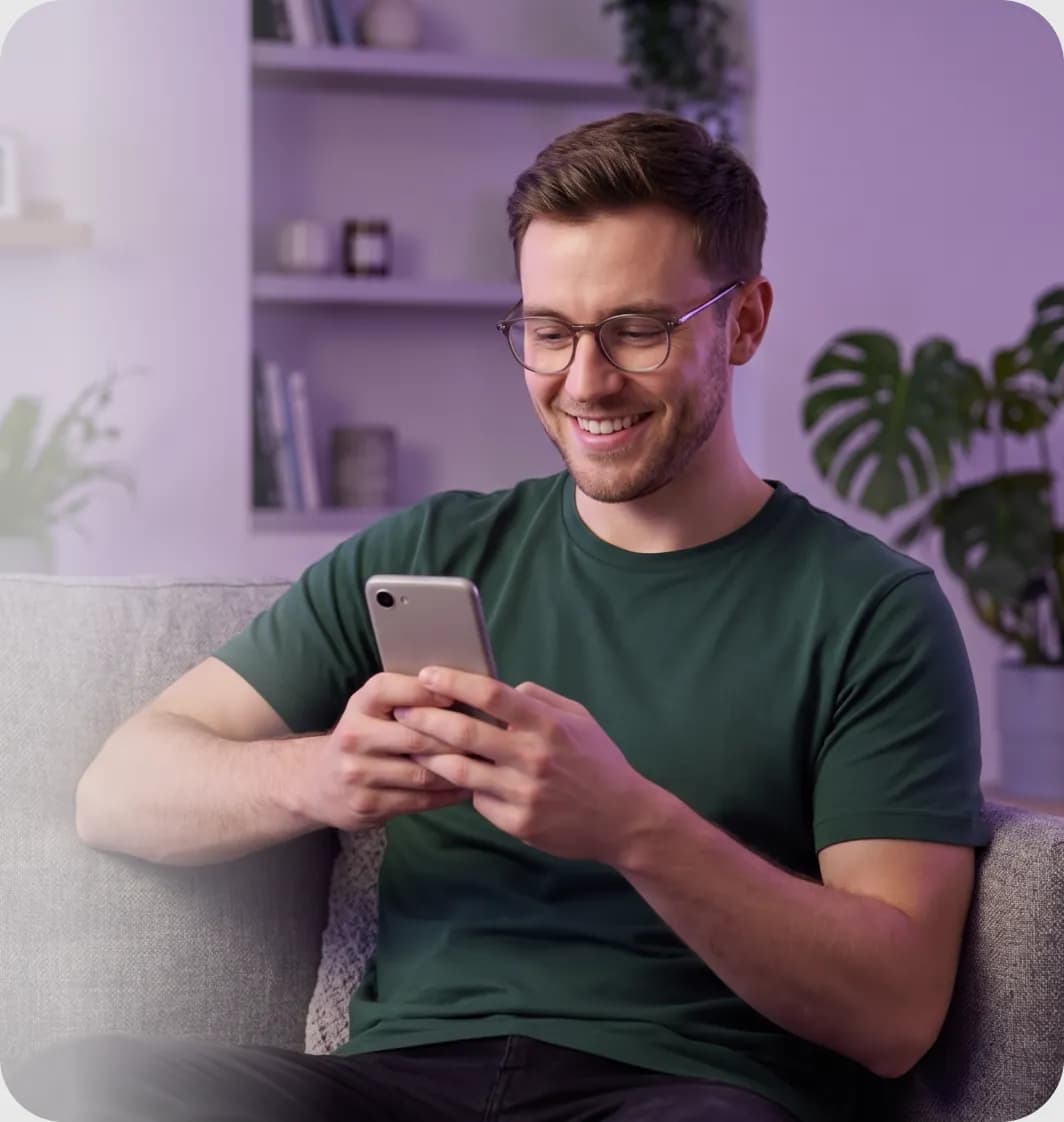 Kanapén ülő, mosolygó, szemüveges férfi a mobiltelefonját nézi