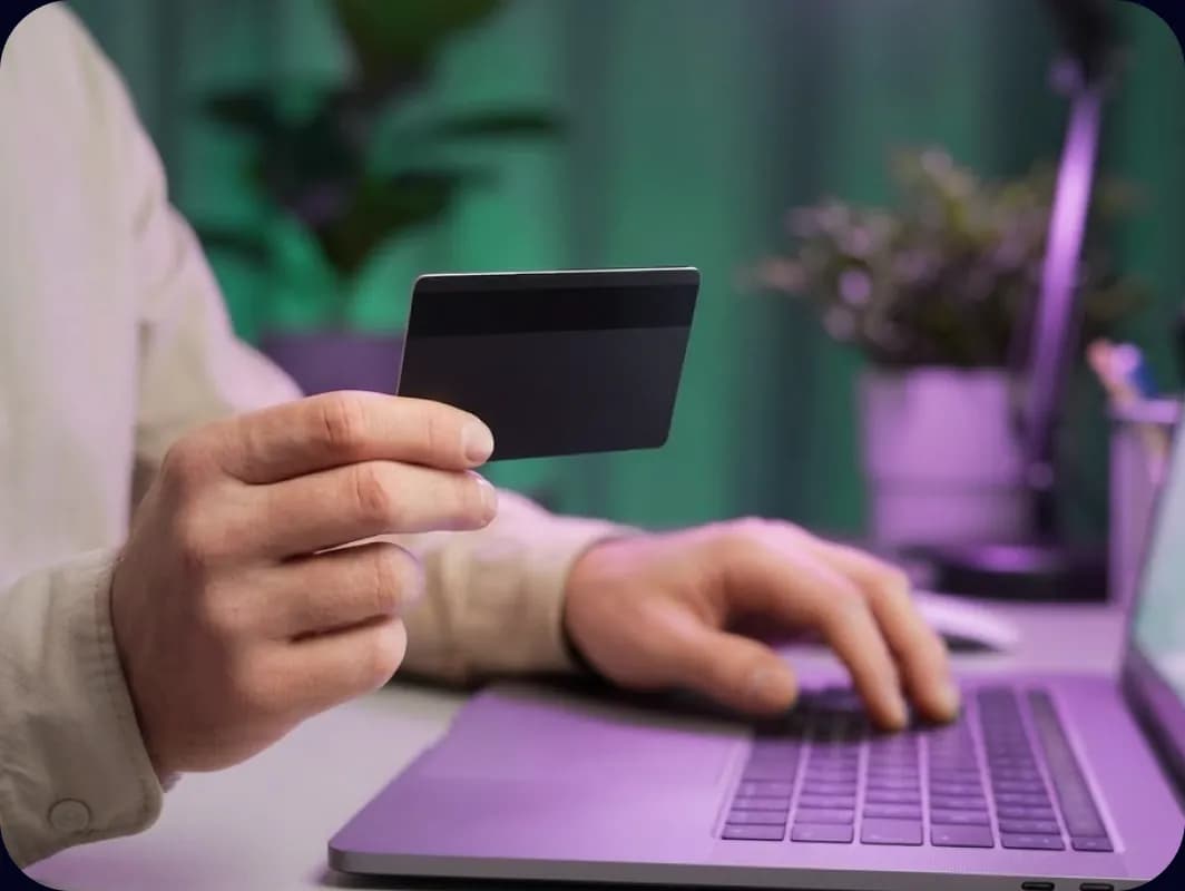 Laptop előtt ülő ember bankkártyát tart a kezében, a háttérben szobanövények