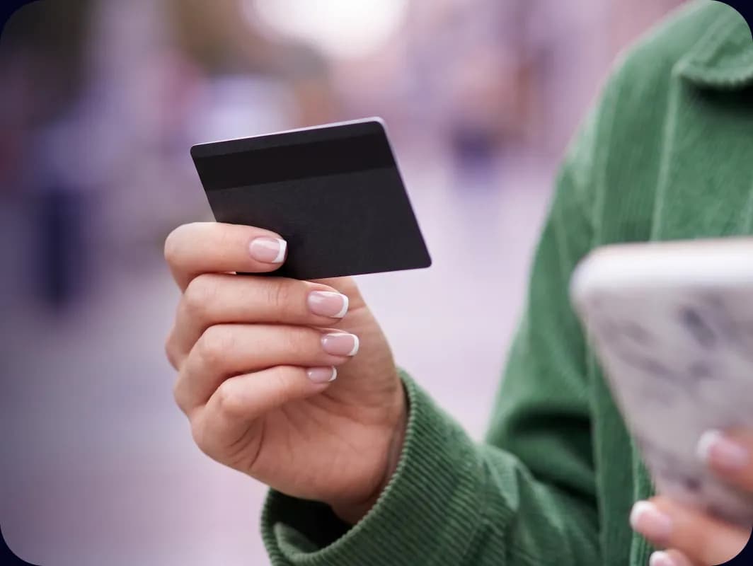 Zöld kabátos nő az utcán, egyik kezében bankkártyát, másikban a telefonját tartja